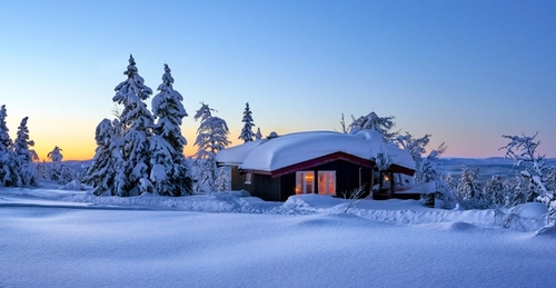 Bilde av hytte med lys i vinduer og snø på taket i vinterlandskap og snødekte trær.