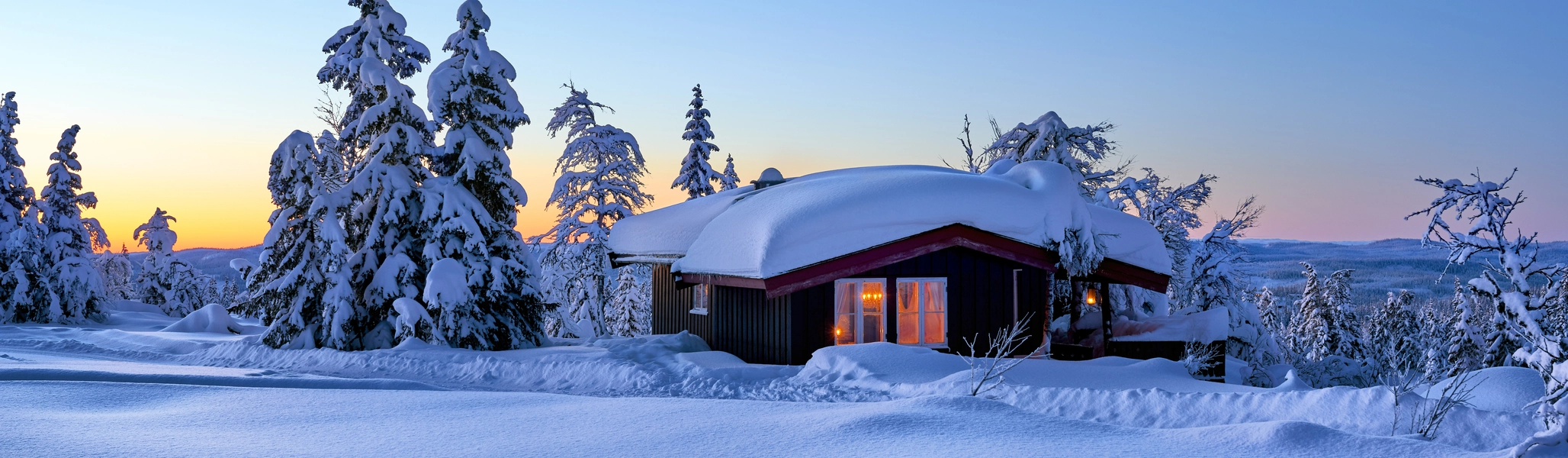 Bilde av hytte med lys i vinduer og snø på taket i vinterlandskap og snødekte trær.