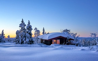 Bilde av hytte med lys i vinduer og snø på taket i vinterlandskap og snødekte trær.