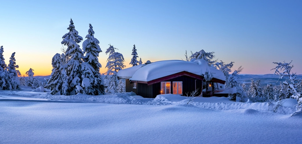 Bilde av hytte med lys i vinduer og snø på taket i vinterlandskap og snødekte trær.